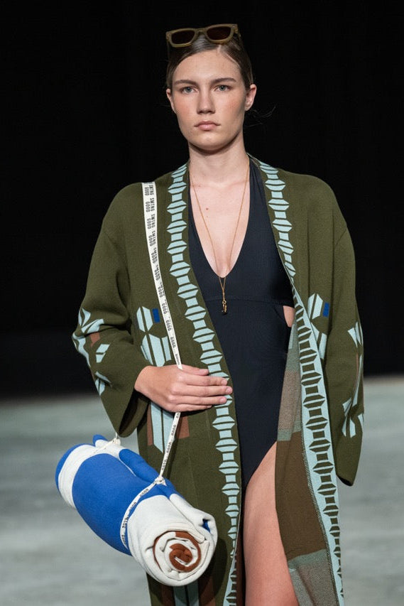 Model on a runway wearing a green patterned cardigan over a black swimsuit, holding a blue and white bag.